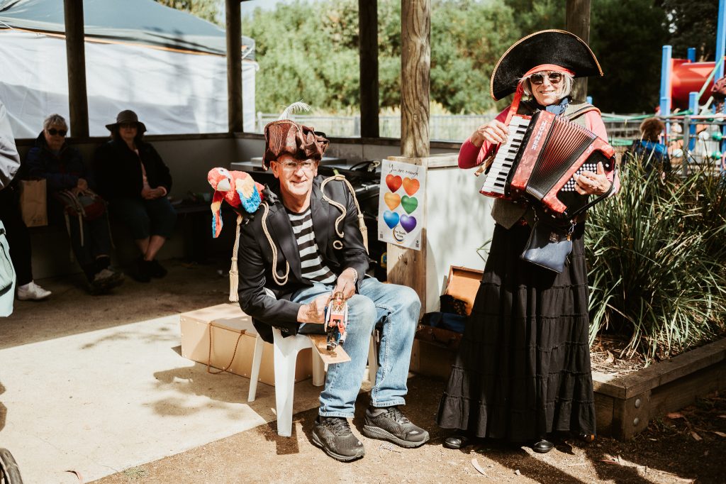 Two adults dressed as pirates, one sitting and playing a tambourine with a parrot on their shoulder, and the other standing while playing an accordion, smile at an outdoor event with people and a playground in the background.