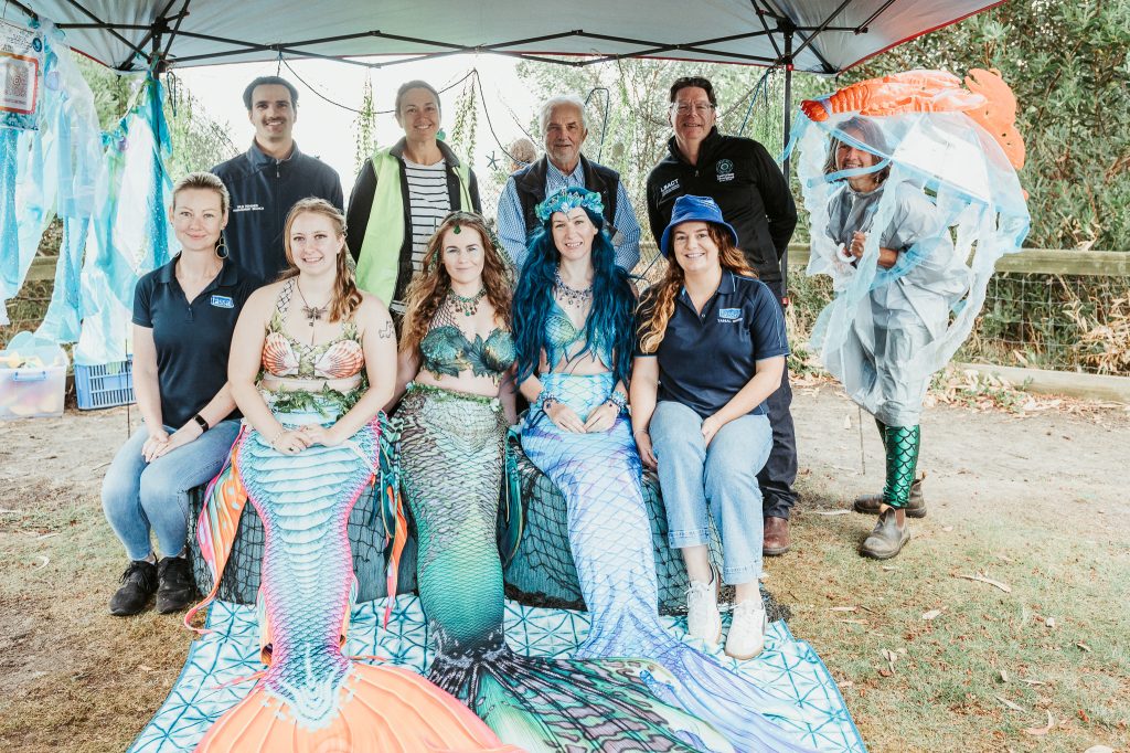 A group of people pose under a canopy, with three women dressed as mermaids sitting in front and others standing or kneeling behind them. The setting appears outdoors with greenery and festival decorations.