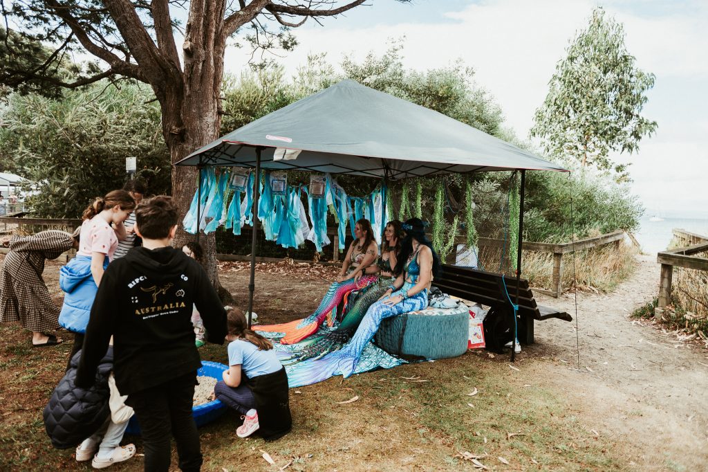People gather around a tented booth by the water, where two women dressed as mermaids sit on a bench. Blue costumes hang from the tent, and children interact nearby in a natural, outdoor setting.