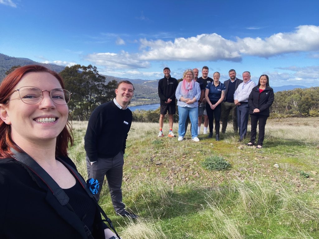 A group of nine people stand and smile on a grassy hillside with trees, a lake, and mountains in the background under a partly cloudy blue sky. One person in the foreground is taking a selfie.