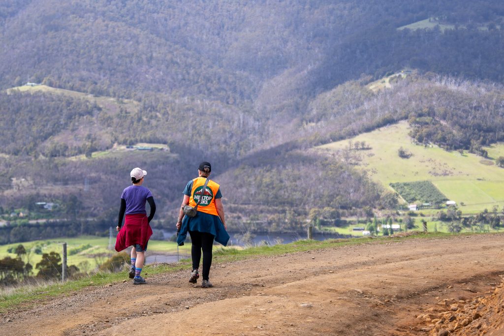 Two people are walking on a dirt path in a hilly landscape, wearing colorful clothes and backpacks. Green fields, scattered houses, and forested hills are visible in the background.