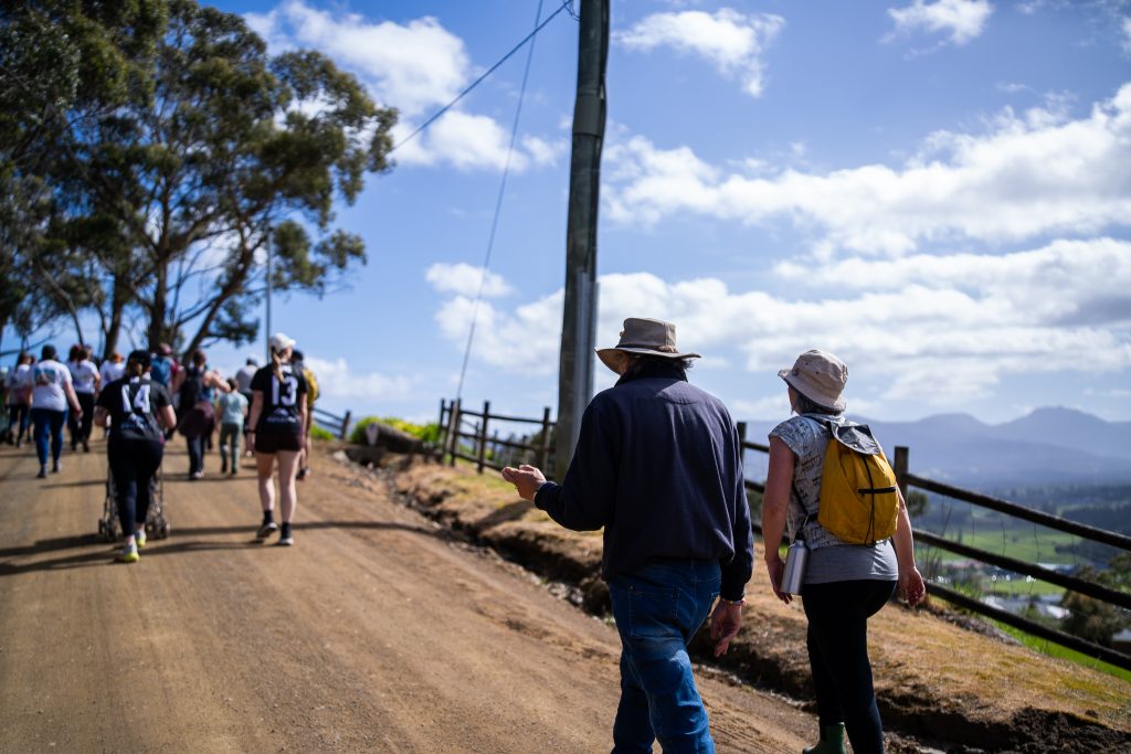 A group of people walk up a dirt road on a sunny day, with some wearing hats and backpacks. Trees, a wooden fence, and mountains are visible in the background under a partly cloudy sky.