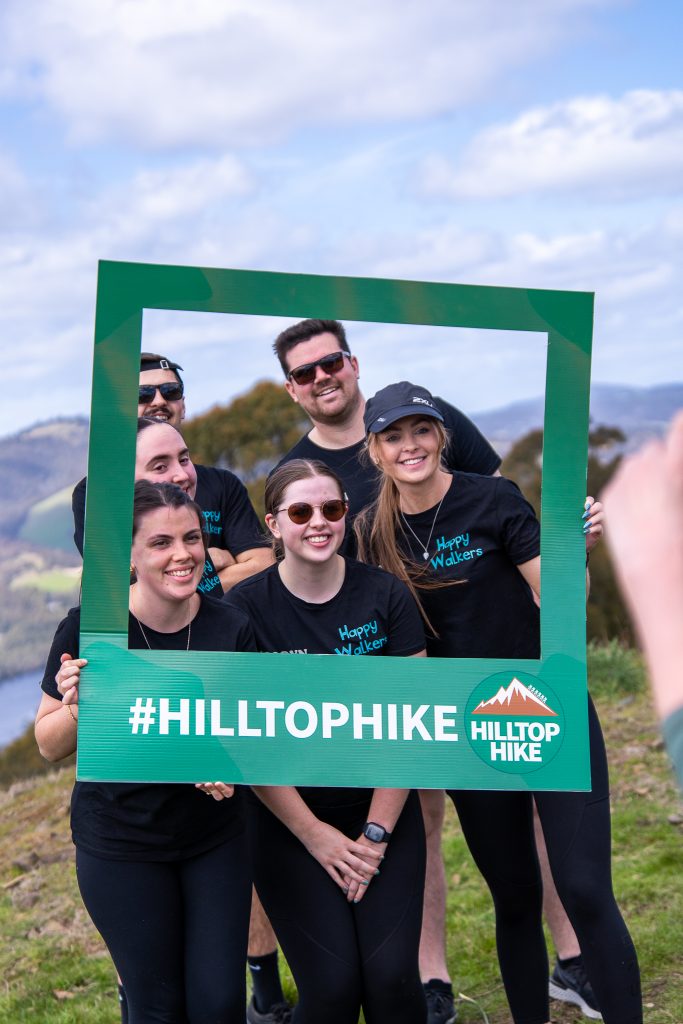 Six smiling people wearing black “Happy Hikers” shirts pose together on a grassy hill, holding a green frame labeled “#HILLTOPHIKE HILLTOP HIKE,” with scenic mountains and sky in the background.