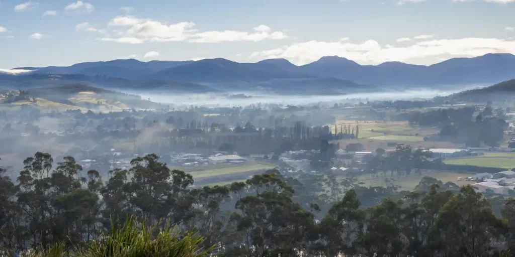 A scenic landscape of a rural valley with mist hovering above fields, houses, and scattered trees, surrounded by rolling hills and silhouetted mountain ranges under a partly cloudy sky.