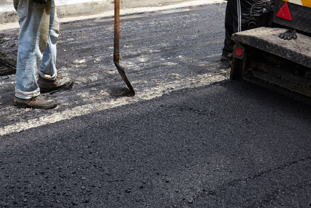 A construction worker stands on a road, holding a shovel while spreading fresh asphalt. Part of a paving machine is visible on the right side of the image. Only the lower bodies of the workers can be seen.