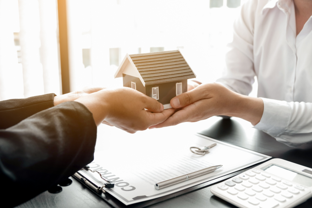 Two people exchange a model house over a desk with contract papers, a pen, a calculator, and a key, symbolizing a real estate transaction or home purchase agreement.