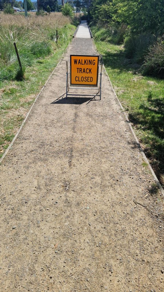 A gravel walking path is blocked by a yellow sign that reads WALKING TRACK CLOSED. The path stretches into the distance, surrounded by grass and trees on both sides.