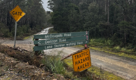 A muddy dirt road in a forest with weathered green road signs pointing to McDougalls Road, Denison Road, and Lake Skinner walking track, alongside warning signs for a gate and traffic hazard ahead.