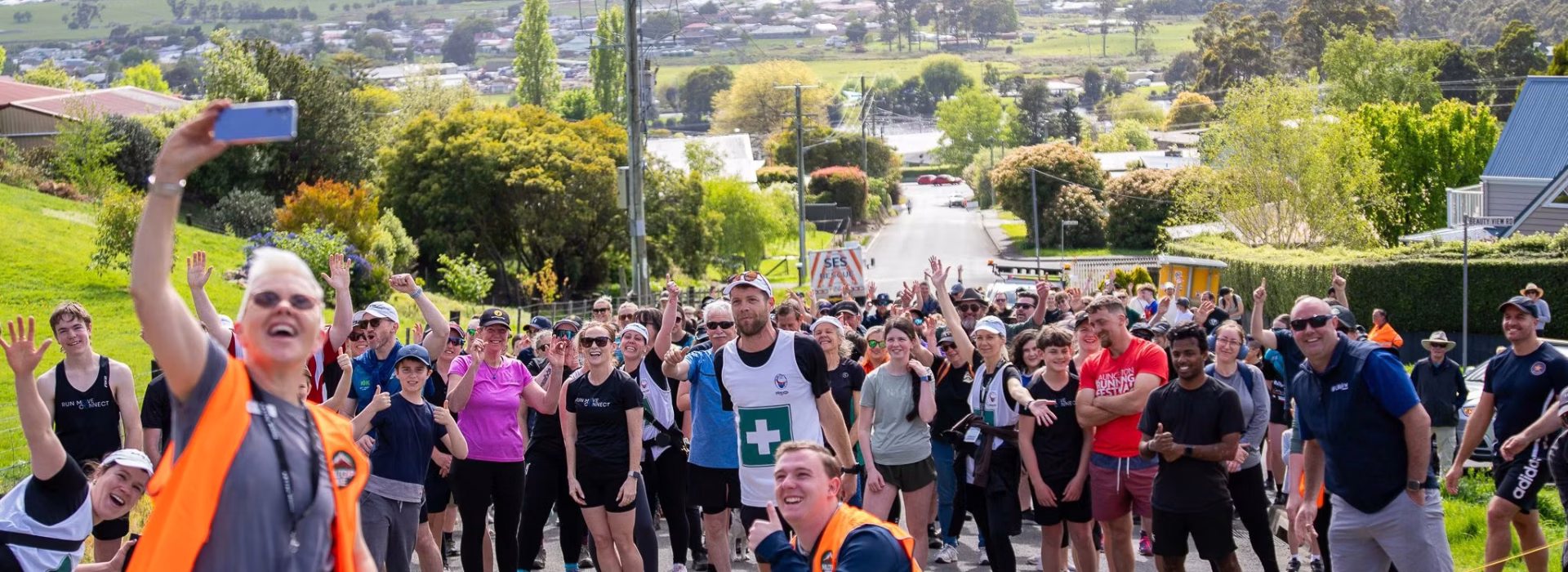 A large group of people in athletic wear gather on a sloped street outdoors, posing and smiling for a group selfie taken by a woman in an orange safety vest. Mountains and houses are visible in the background.