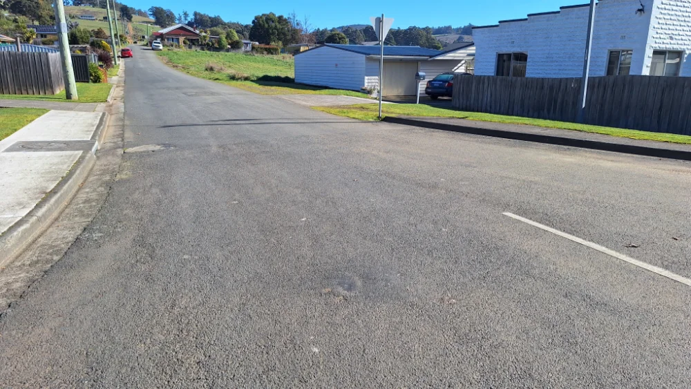 A quiet suburban street with houses, a grassy hill, and a parked car. The road is empty, and theres a sidewalk on the left side. The sky is clear and sunny.