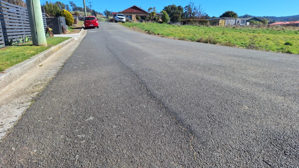 A quiet suburban street with a red car parked on the left, a house at the end of the road, green grass on the right, and clear blue sky overhead.