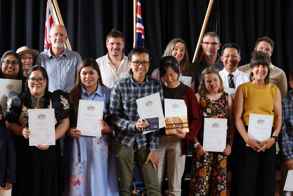 A diverse group of people stands smiling and holding certificates in front of three Australian flags, posing together on a stage with a dark curtain backdrop.