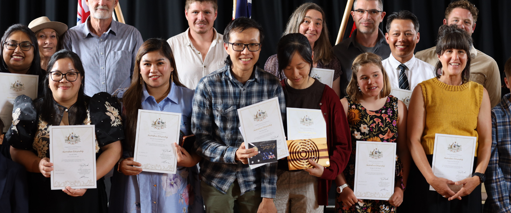 A diverse group of people stands smiling and holding certificates in front of three Australian flags, posing together on a stage with a dark curtain backdrop.