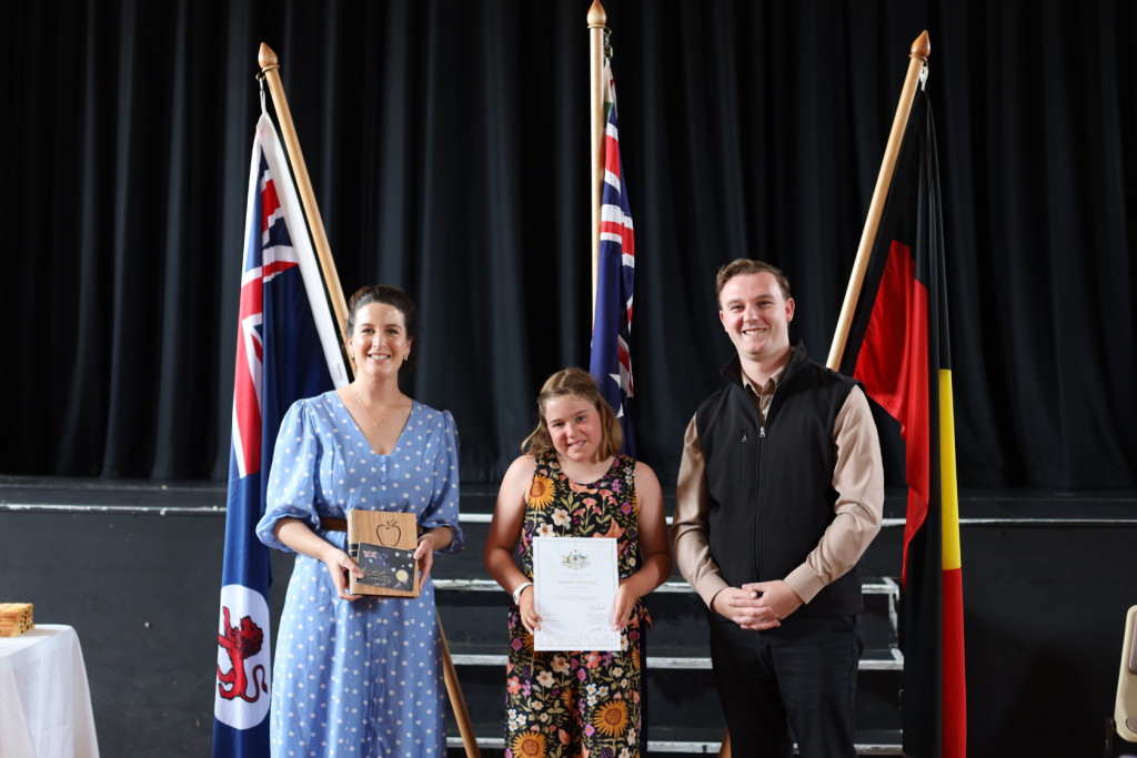 Three people stand smiling in front of three flags on a stage. The person in the middle holds a certificate, the person on the left holds a wooden award, and the person on the right stands with hands clasped.
