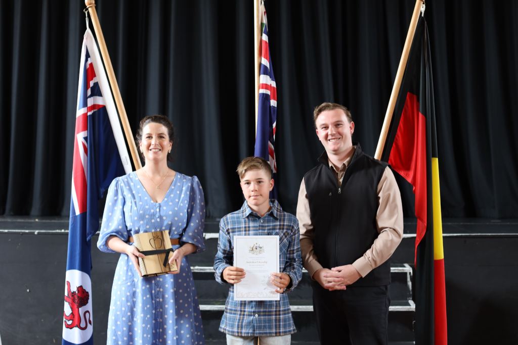 Three people stand indoors in front of three flags. The woman on the left holds a gift, the boy in the center holds a certificate, and the man on the right smiles. They are dressed smartly, likely at an award ceremony.