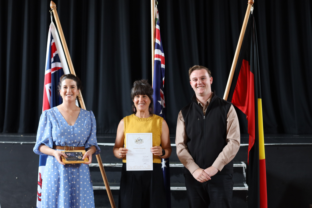 Three people stand in front of three flags on a stage. The woman in the center holds a certificate, the woman on the left holds a box, and the man on the right stands with hands clasped. All are smiling.