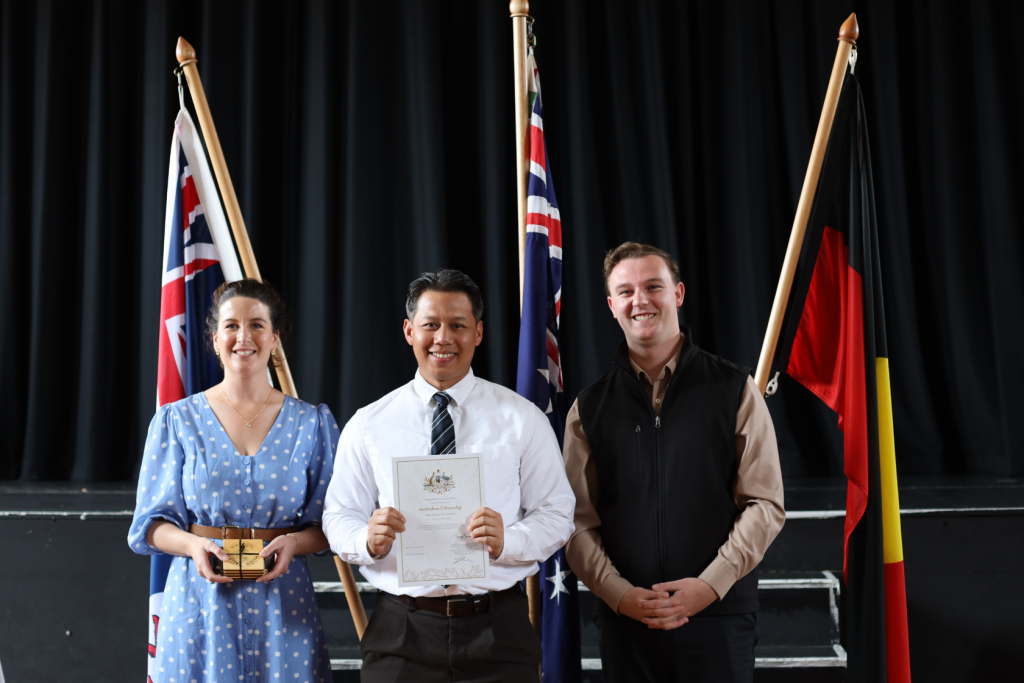 Three people stand smiling in front of three flags (including Australian and Aboriginal flags). The person in the middle holds a certificate, the person on the left holds a small box, and the person on the right stands with hands clasped.
