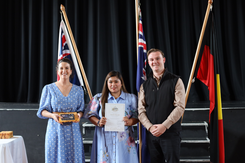 Three people stand indoors in front of three flags. The woman on the left holds a wooden plaque, the woman in the center holds a certificate, and the man on the right stands with his hands clasped, all smiling at the camera.