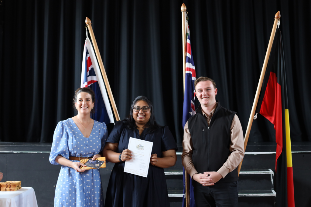 Three people stand in front of Australian and Aboriginal flags. The person in the center holds a certificate and smiles, while the other two stand on either side, also smiling.