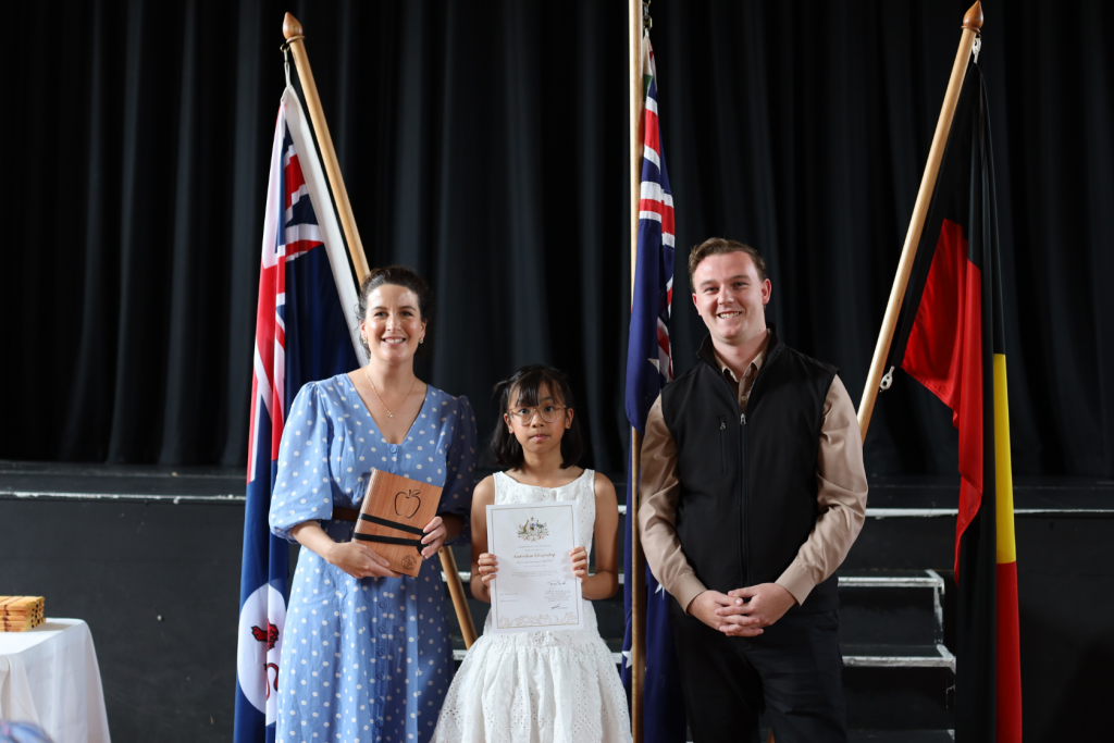 Three people stand in front of three flags on a stage. A girl in a white dress holds a certificate, flanked by a smiling woman in a blue dress with a plaque and a man in a dark vest and shirt.