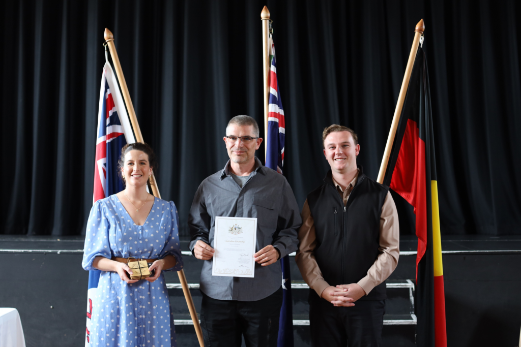 Three people stand indoors in front of three flags, smiling at the camera. The person in the middle holds a certificate, the person on the left holds a small trophy, and the person on the right stands with hands clasped.