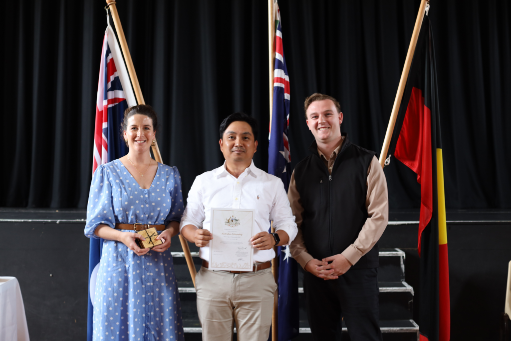 Three people stand indoors in front of two Australian flags and an Aboriginal flag. The person in the middle holds a certificate, while the others smile at the camera. The background is a black curtain with steps.