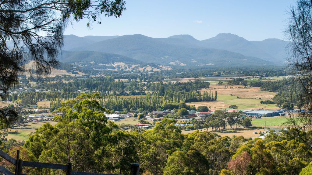 A scenic view of a rural valley with scattered houses and farms, green trees in the foreground, and layered blue mountains in the distance under a clear sky.