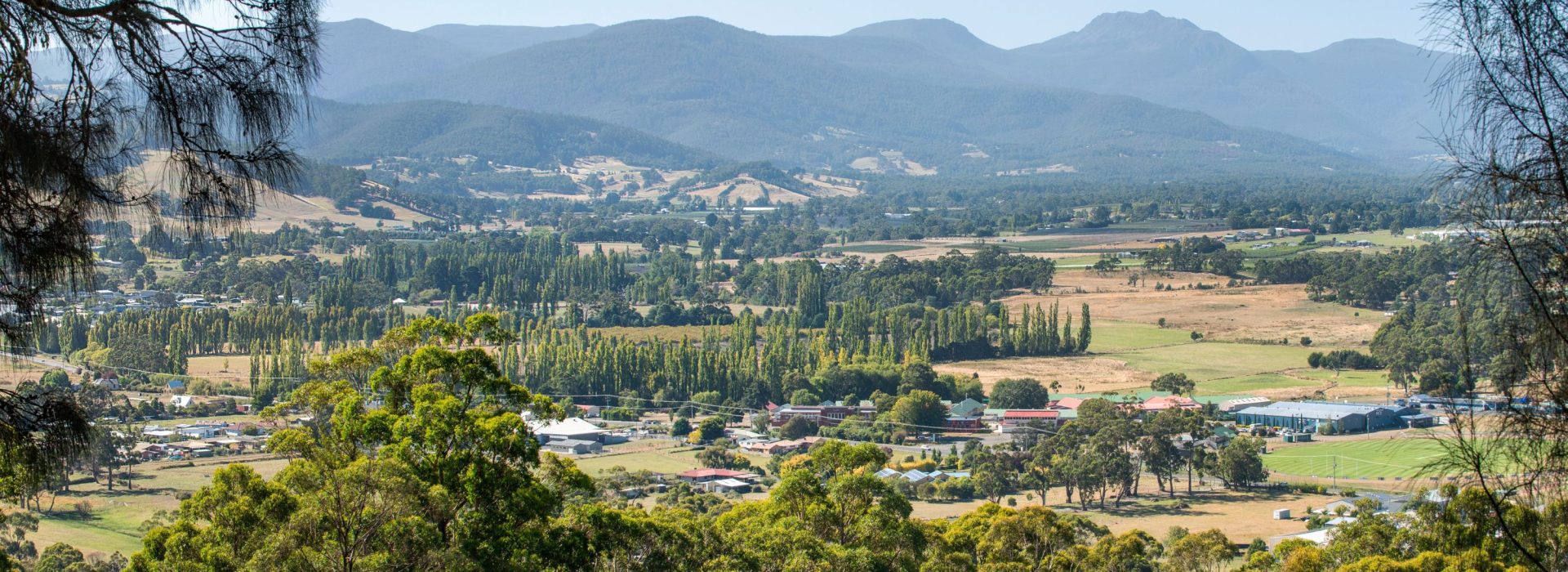 A scenic view of a rural valley with scattered houses and farms, green trees in the foreground, and layered blue mountains in the distance under a clear sky.
