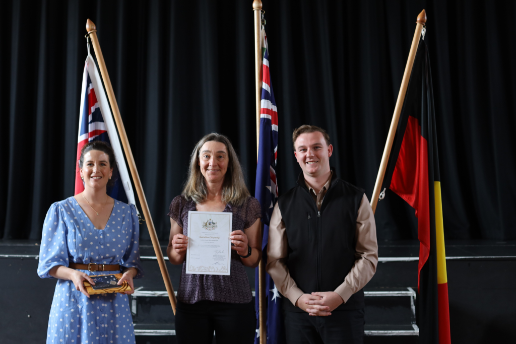 Three people stand smiling indoors between three flags, with the person in the middle holding a certificate. The background features a stage with dark curtains. The person on the left holds a small object.