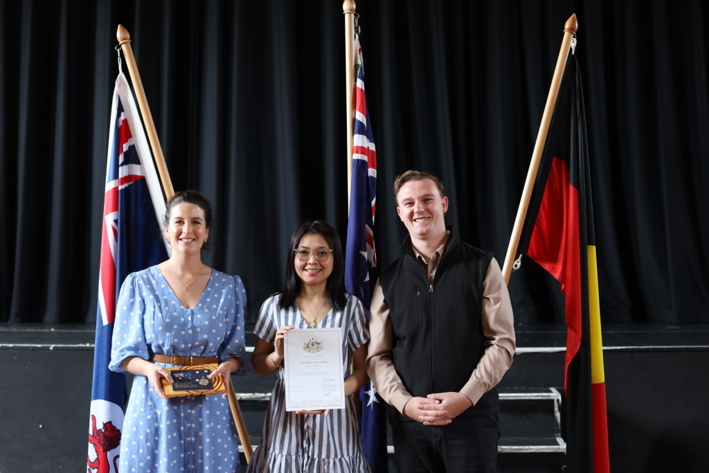 Three people stand smiling in front of three flags, including the Australian and Aboriginal flags. The person in the middle holds a certificate; the person on the left holds a small award. They are positioned on a stage with a black curtain backdrop.