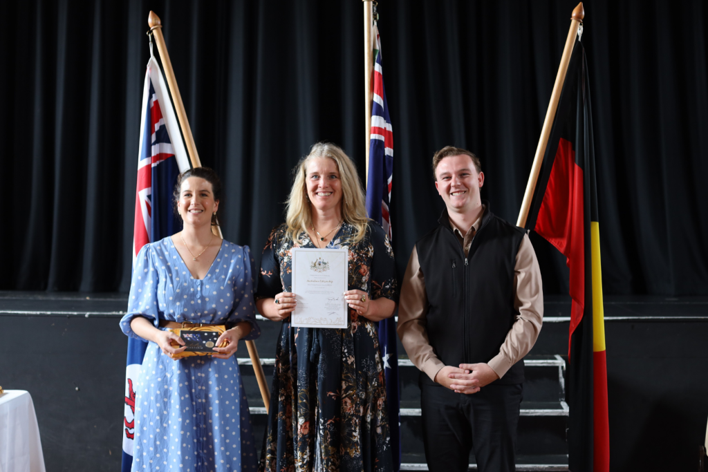 Three people stand smiling in front of three flags on a stage. The woman in the center holds a certificate, the woman on the left holds a small box, and the man on the right stands with his hands clasped.