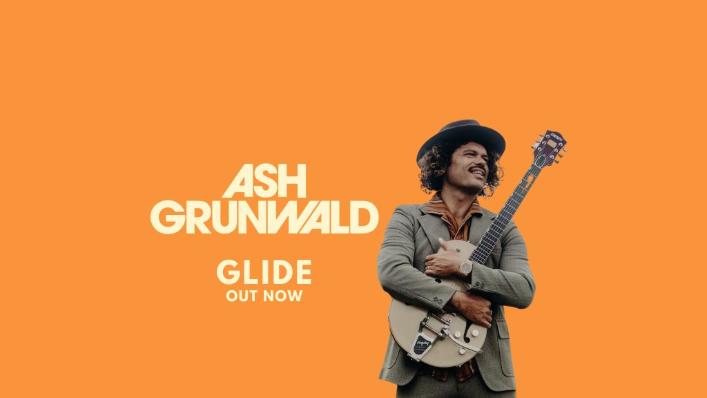 A man in a suit and hat hugs an electric guitar and smiles. The orange background features bold white text reading ASH GRUNWALD and below it, GLIDE OUT NOW.