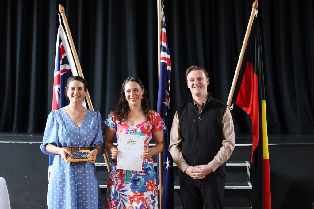 Three people stand smiling in front of Australian and Aboriginal flags. The woman on the left holds a small box, the woman in the middle holds a certificate, and the man on the right stands with his hands together.