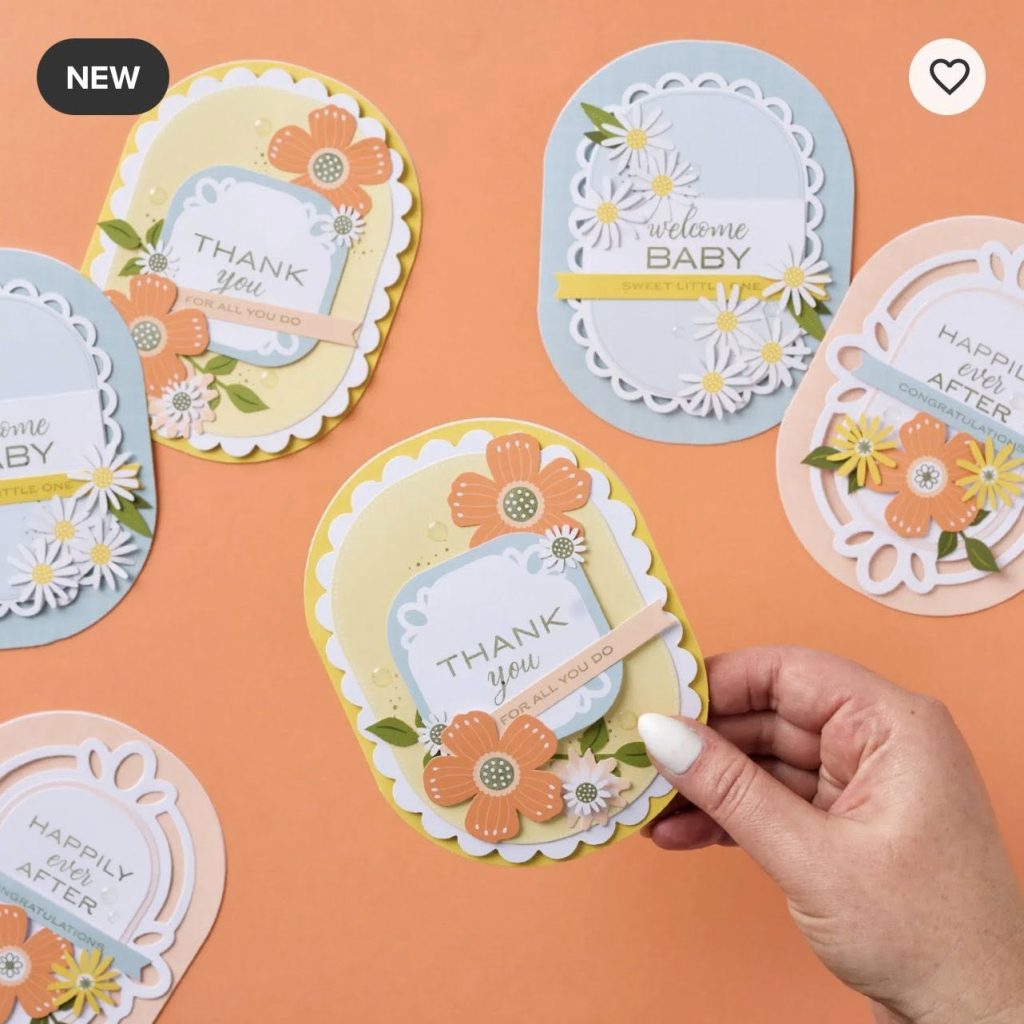 A hand holds a decorative card reading “Thank you for all you do” with flowers and scalloped edges. Similar pastel cards with messages like “welcome baby” and “happily ever after” are spread on an orange surface.