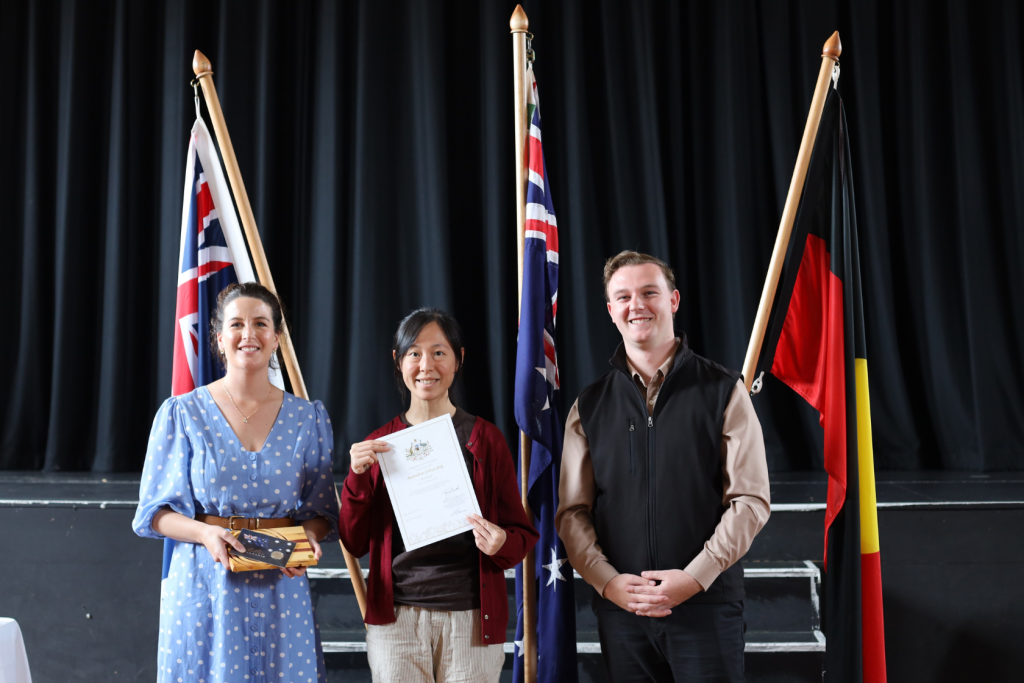 Three people stand indoors in front of three flags, with one person in the center holding a certificate and smiling. The person on the left holds a box, and all appear to be at a formal event.