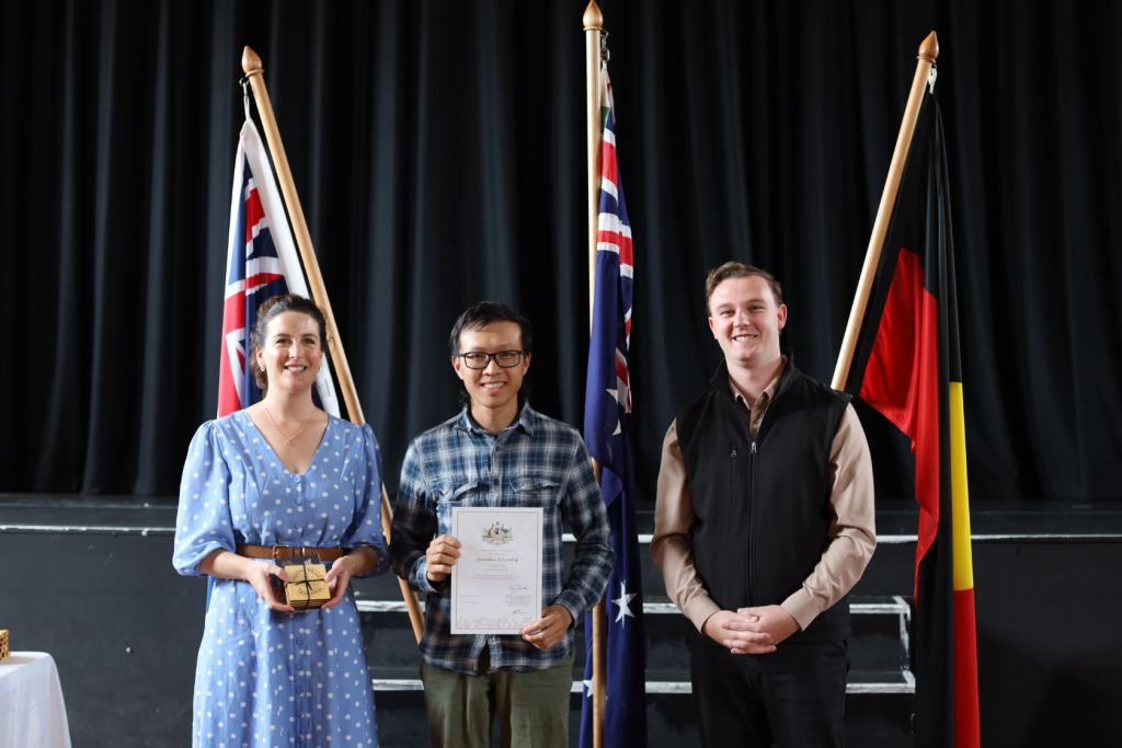 Three people stand smiling indoors in front of three flags, including the Australian flag. The central person holds a certificate, the left person holds a gift, and the right person stands with hands clasped.