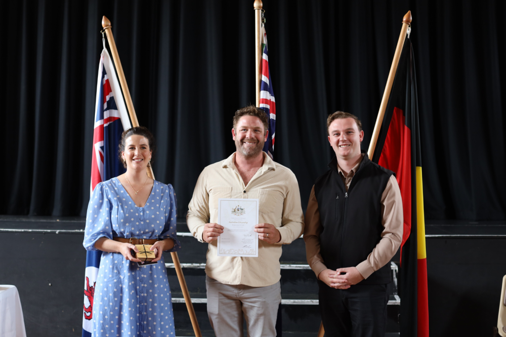 Three people stand smiling indoors in front of Australian and Aboriginal flags. The person in the center holds a certificate, while the person on the left holds a small box. All are dressed formally. A black curtain is in the background.