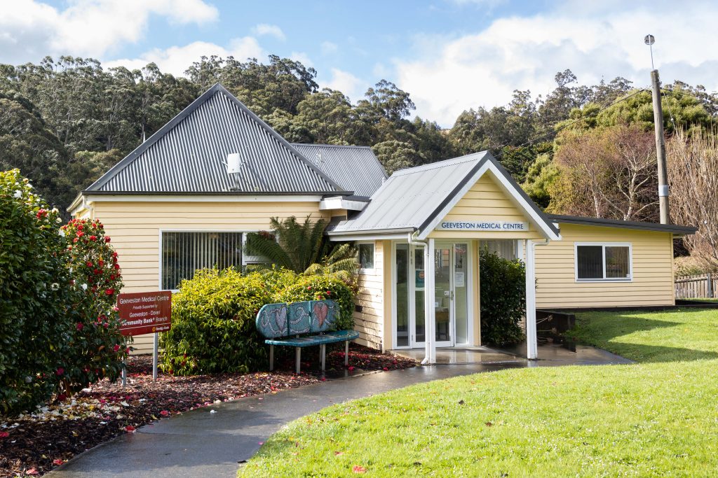A small, single-story yellow building with a metal roof and a sign reading Geeveston Medical Centre stands surrounded by greenery, trees, and a garden bench near the entrance on a clear day.