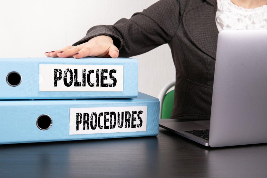 A person in business attire sits at a desk with a laptop, resting their hand on two large blue binders labeled POLICIES and PROCEDURES.