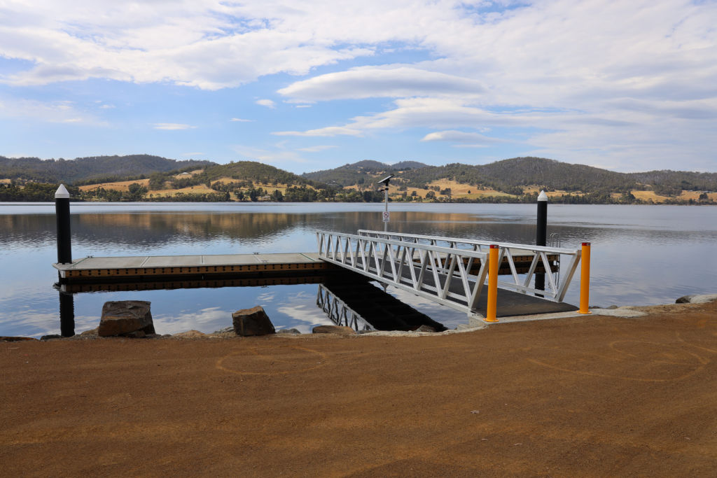 A floating dock with metal railings extends over a calm lake, reflecting clouds and surrounding hills. The shore is covered in brown dirt, and the landscape features green and golden hills under a partly cloudy sky.