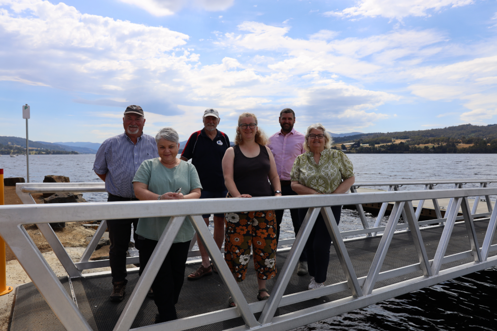 Six adults stand on a metal dock by a lake, with hills and partly cloudy sky in the background. Some are smiling, and all are casually dressed. The water and landscape create a scenic, relaxed atmosphere.