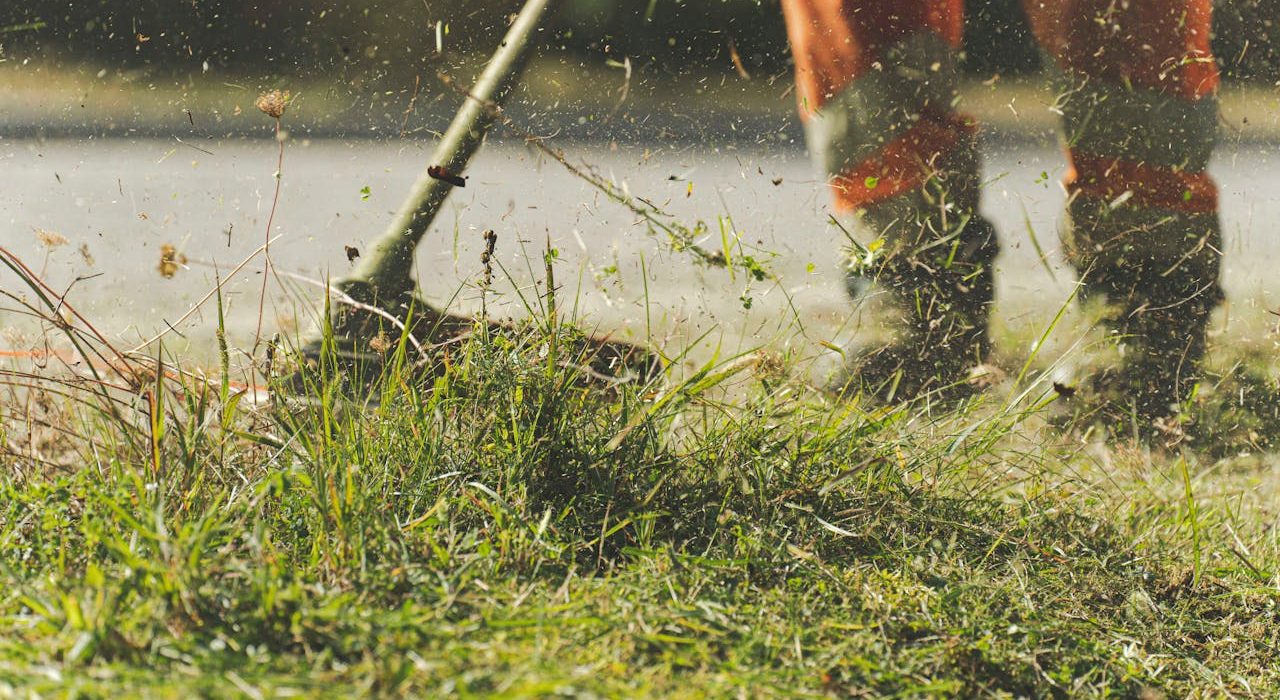 Close-up of a person wearing orange pants using a string trimmer to cut grass, with blades and debris flying in the air. The persons upper body is not visible.