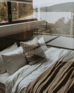 A cozy bed with white sheets, pillows, and a brown throw blanket sits by a large window overlooking a scenic lake and forested hills. The room has a modern, minimalist style.