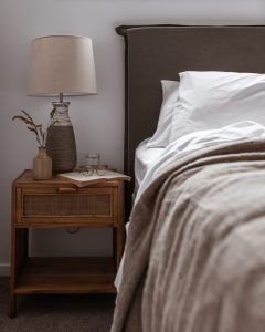 A cozy bedside scene with a wooden nightstand holding a ceramic lamp, a small vase with dried grass, a glass of water, eyeglasses, and an open book next to a neatly made bed with white pillows and beige bedding.