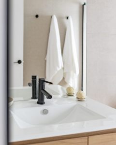 A modern bathroom sink with a black faucet, two round beige soap bars on the counter, and two white towels hanging on hooks in the background, reflected in a large mirror.