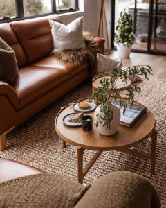 A cozy living room features a brown leather sofa with pillows and a fur throw, a round wooden coffee table with a white vase holding green leaves, snacks, and books, all on a woven rug beside a window and houseplant.