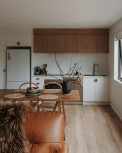 A modern kitchen and dining area with wood floors, white cabinets, a stainless steel dishwasher, a refrigerator, and a wooden dining table set for three. A vase with branches decorates the table; natural light fills the room.