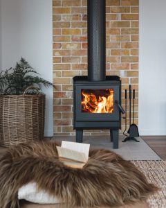 A black wood-burning stove with a fire inside stands against a brick wall. In front, a fluffy brown rug holds an open book. A wicker basket with a plant and fireplace tools are nearby, creating a cozy living space.
