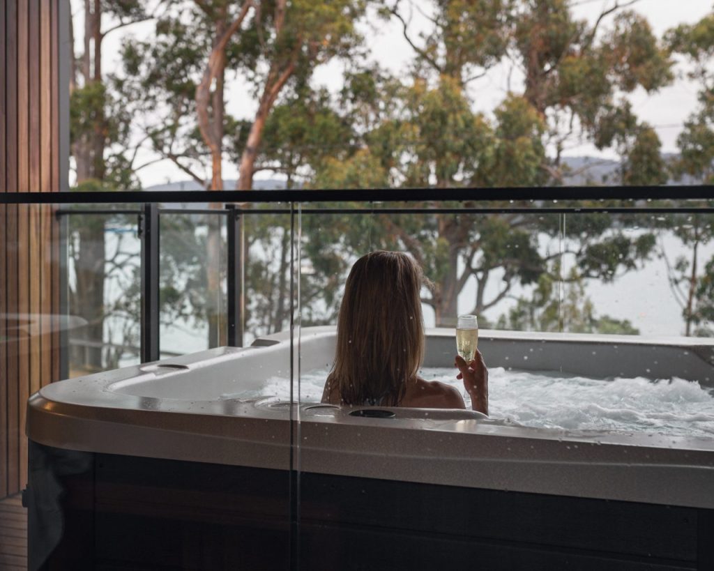 A person with long hair relaxes in an outdoor hot tub on a balcony, holding a glass of champagne and gazing at trees and water in the background.
