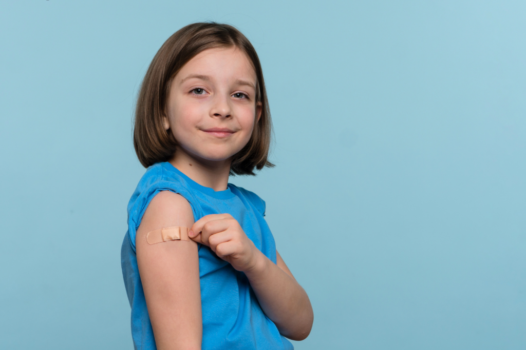 A young girl in a blue shirt smiles gently and shows her upper arm with a bandage, indicating she just received a vaccination. The background is a plain light blue.